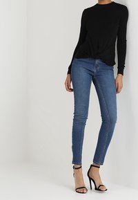 Femme portant un haut noir à manches longues, un jean skinny bleu et des sandales à talons hauts noires, se tenant devant un mur blanc uni.