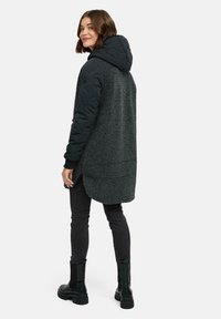 Mörkgrå jacka med en quiltad huva och ärmar, som har en texturerad, fluffig kropp, sidoflikar och svarta åtsittande byxor med kraftiga stövlar.