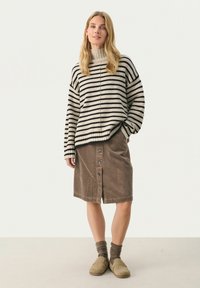 Übergroßer schwarz-weißer gestreifter Pullover mit Rollkragen, kombiniert mit einem braunen Cordrock mit Knöpfen auf der Vorderseite. Braune Holzclogs und Socken.