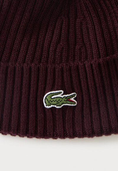Bonnet tricoté violet foncé avec texture côtelée et petit logo crocodile vert brodé sur le revers.