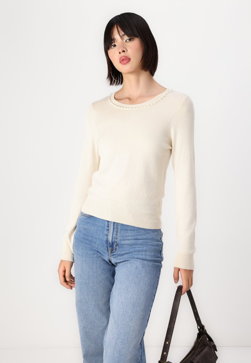 Maglione crema con maniche lunghe, scollatura rotonda accentuata da piccole perline, abbinato a jeans azzurri chiari e una borsa scura. Texture morbida.