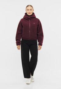 Burgundin fleece-takki, jossa on vetoketju edessä, iso huppu ja kaksi etutaskua, yhdistetty mustiin leveälahkeisiin housuihin ja valkoisiin lenkkarit.