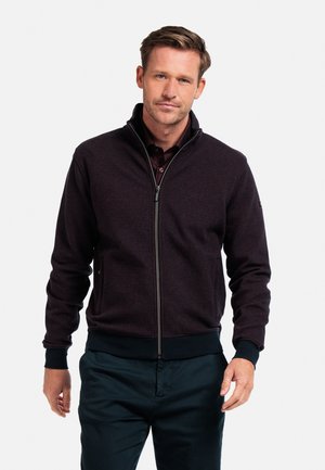 Herren Zip-Jacke in dunkelmaroon, mit einem strukturierten Stoff, hohem Kragen und gerippten Bündchen. Kombiniert mit navyfarbigen Hosen.