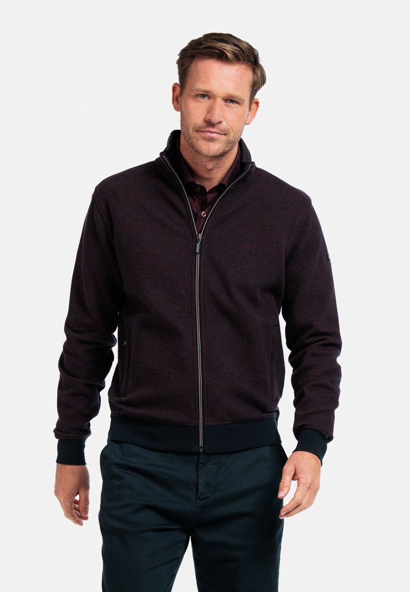Heren zip-up jas in donker bordeaux, met een gestructureerde stof, hoge kraag en geribbelde manchetten. Gecombineerd met marineblauwe broeken.