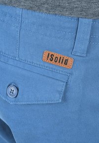 Shorts en coton bleu avec une poche arrière à fermeture par bouton et une étiquette en cuir marron embossée avec "!Solid". Subtils détails de texture et de couture.