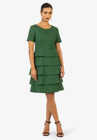 Grünes, kurzärmliges Kleid mit Rundhalsausschnitt und geschichteten Volantdesign, aus strukturierter Stoffqualität, kombiniert mit schwarzen Absatzsandalen.