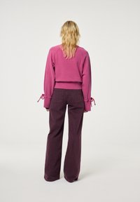 Afgeknotte roze sweatshirt met gathered mouwen en strikdetails, gecombineerd met donker bordeaux brede broek, gezien vanuit de achterkant.