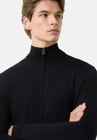 Schwarzer, enganliegender Reißverschluss-Pullover mit hohem Kragen, Strickstruktur und einem kleinen silbernen Reißverschlussdetail am Hals. Minimalistisches und elegantes Design.