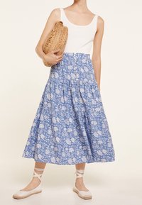 Jupe midi imprimée fleuri bleu avec des volants froncés, assortie à un débardeur blanc et une pochette en paille ; modèle portant des chaussures claires avec des lanières à la cheville.