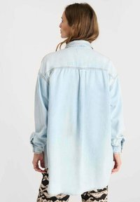 Lichtblauwe denim shirt met lange mouwen, ontspannen pasvorm en knoopsluiting kraag. Heeft een steek achter en geleidelijke vervaging voor textuur.