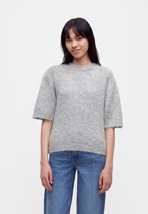 Ung kvinde med glat mørkt hår iført en løs grå kortærmet sweater og blå jeans, stående mod en ensfarvet hvid baggrund.