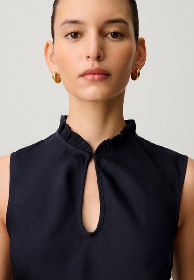 Joven mujer con pendientes de aro dorados y una blusa negra sin mangas con cuello fruncido y escote en forma de ojo de cerradura.