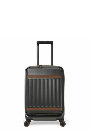 Musta kovakuorinen matkalaukku, jossa on sisäänvedettävä kahva, ruskeat korosteraidat, edessä vetoketjullinen lokero ja neljä pyörivää pyörää, pystyssä seisova.