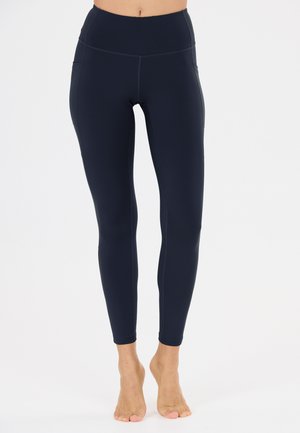 Kvinde iført sorte, højtaljede leggings med sidelommer, står barfodet mod en ensfarvet hvid baggrund.
