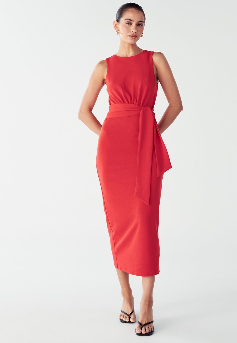 Rotes ärmelloses Kleid mit einem figurbetonten Oberteil, breitem Taillenband und einer gebundenen Schleife. Glatter, dehnbarer Stoff, knielang, kombiniert mit schwarzen Sandalen.