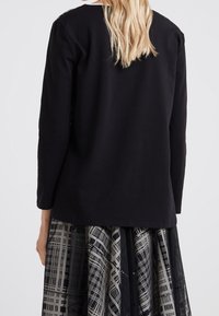 Blonde-haired person wearing a black long-sleeve top and a black-and-white patterned skirt, shown from the back against a plain background.