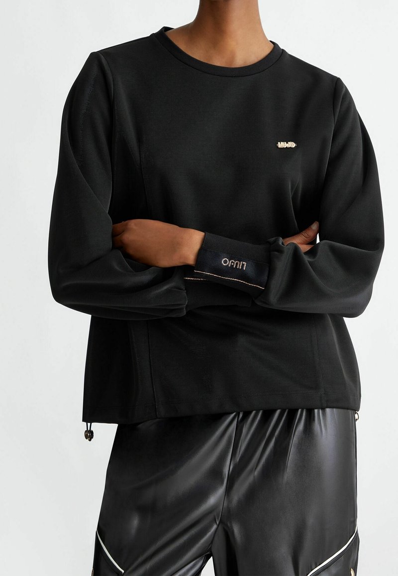 Personne portant un sweat-shirt noir à manches longues avec un petit logo doré sur la poitrine et un pantalon en cuir noir avec une bordure blanche aux genoux.