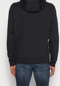 Sudadera negra con capucha de textura suave, de mangas largas y con un bolsillo tipo canguro. Combinada con unos vaqueros de mezclilla azul oscuro con bolsillos traseros.