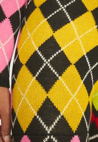 Peça de vestuário tricotada em múltiplas cores, com um padrão em diamante em rosa, amarelo, preto e verde, com detalhes em branco e tecido texturizado.