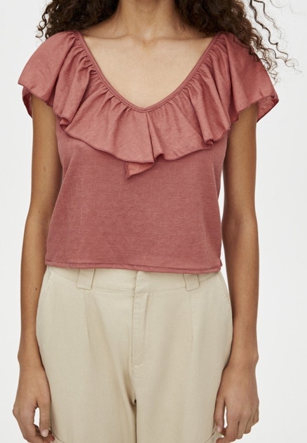 Femme portant un top rose poudré à manches courtes à volants et un pantalon beige taille haute, debout devant un fond blanc uni.