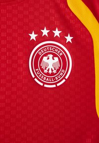 Κόκκινη αθλητική φανέλα με στρογγυλό λευκό λογότυπο που απεικονίζει έναν αετό και πέντε αστέρια. Κίτρινες λεπτομέρειες στους ώμους και υφή υφάσματος.