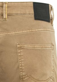 Beige denim broek met een achterzak, gestikte details en een zwart label op de taille. Soepele textuur met subtiele kleurvariaties.