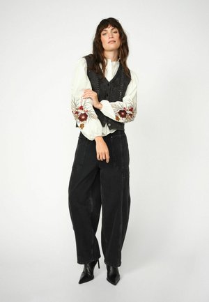 Zwart gebreide vest over een witte blouse met bloemenborduursel op de mouwen, gecombineerd met losse zwarte broeken en zwarte puntige hakken.