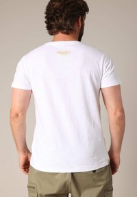 Witte katoenen t-shirt met korte mouwen, voorzien van een klein gouden tekstontwerp op de achterkant. Klassieke ronde hals, zachte textuur, comfortabele pasvorm.