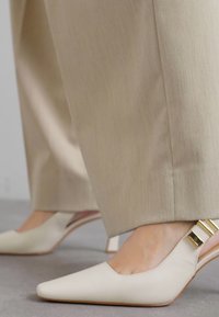 Kreminės odinės aukštakulnės su atvira kulnike, su smailiu kitu ir auksinės spalvos detalėmis bei lygiu paviršiumi. Dėvimos su šviesių spalvų kelnėmis.