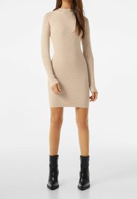 Robe tricotée en beige clair avec une silhouette ajustée, des manches longues et un col montant. Associée à des bottines noires.