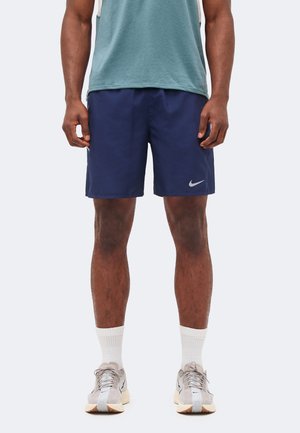 Mężczyzna ubrany w granatowe spodenki Nike, białe skarpetki i beżowe buty sportowe stoi na jednolitym tle.