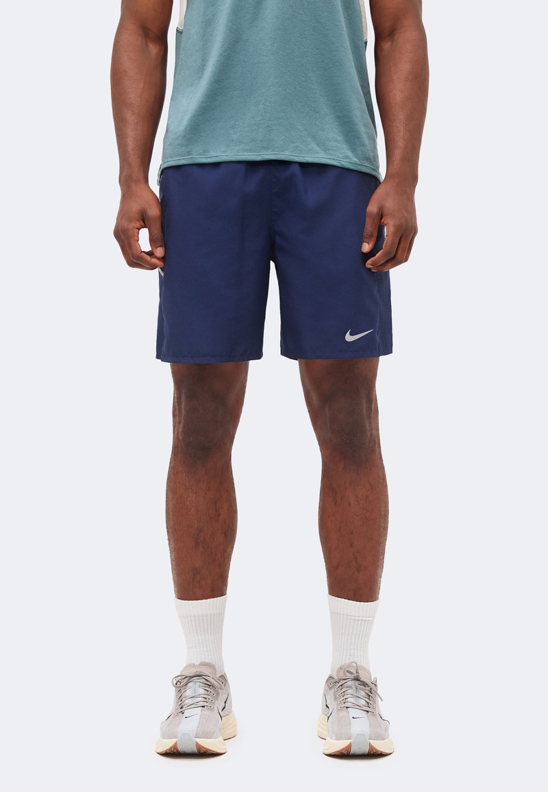 Άνδρας που φοράει σκούρο μπλε σορτς Nike, λευκές κάλτσες και μπεζ αθλητικά παπούτσια, στέκεται μπροστά σε σκονισμένο φόντο.