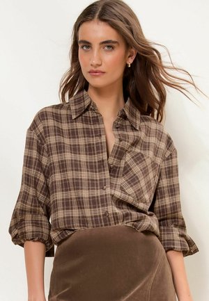 Woman with long brown hair wearing gold earrings, a brown plaid button-up shirt, and a brown skirt against a plain light background.