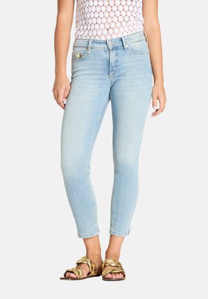 Femme portant un jean skinny court bleu clair avec un haut en crochet blanc et des sandales plates dorées, se tenant devant un fond uni.