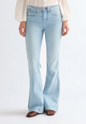 Frau in hellblauen, hoch taillierten Jeans mit Schlag, kombiniert mit braunen, geflochtenen Schuhen und einer weißen Bluse mit langen Ärmeln.