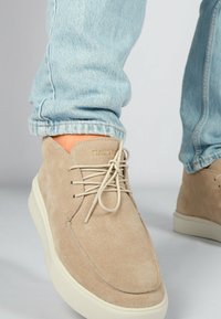 Zapatos de ante beige con cordones crema, punta redonda y una suela blanca gruesa, colocados cerca de unos pantalones vaqueros de tejido ligero con dobladillos acampanados.