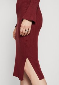 Robe en maille côtelée couleur bordeaux avec manches longues, présentant une fente haute sur le côté et une silhouette ajustée. Texture lisse et extrémités des manches évasées.