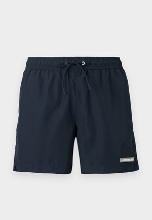 Homme mince à la peau foncée debout pieds nus, portant un short de bain bleu marine foncé avec cordon, regardant vers le bas à sa gauche sur un fond clair uni.