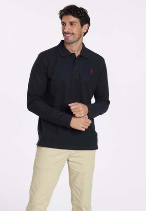 Polo noir à manches longues avec un col, affichant un logo rouge sur la poitrine. Modèle portant un pantalon beige, se tenant devant un fond clair.