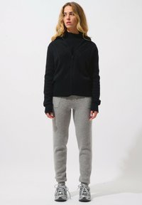 Svart zip-up hoodie med texturerat mönster, tillsammans med ljusgrå träningsbyxor. Modellen har grå sneakers på sig, med händerna vid sidorna i en neutral ställning.