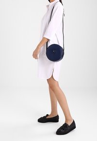 Žena v bílém košilovém šatu s černými mokasíny a námořnickou kulatou crossbody taškou s detaily s přezkou.