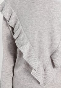 Pull en tricot gris clair à manches longues avec un détail en tricot froncé en diagonale sur le devant et l'épaule.