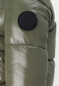 Olive green puffer jacket with a glossy texture, featuring a round black patch with a duck logo labeled "SAVE THE DUCK."