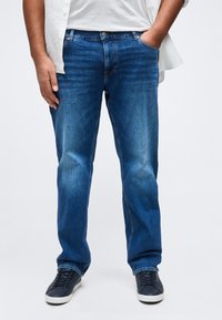 Άνδρας που φοράει μπλε τζιν με ίσια γραμμή, λευκό πουκάμισο και μαύρα αθλητικά παπούτσια, στέκεται με το ένα χέρι στην τσέπη μπροστά σε απλό φόντο.