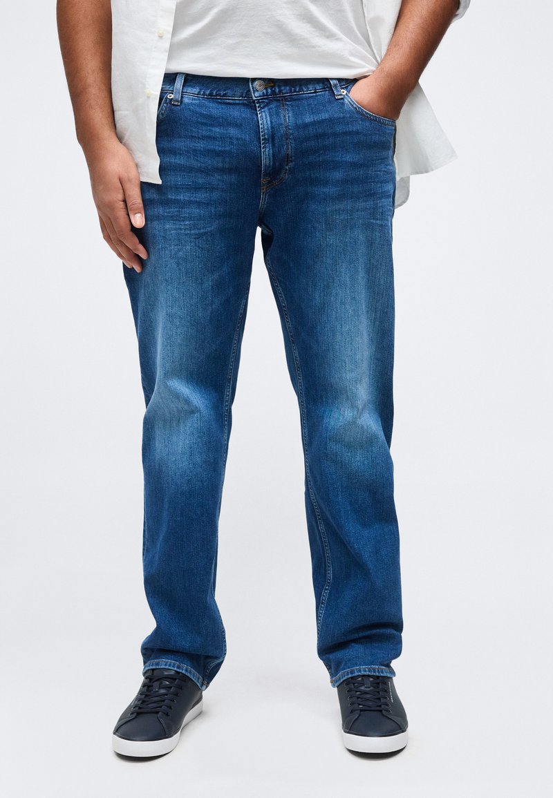 Άνδρας που φοράει μπλε τζιν με ίσια γραμμή, λευκό πουκάμισο και μαύρα αθλητικά παπούτσια, στέκεται με το ένα χέρι στην τσέπη μπροστά σε απλό φόντο.