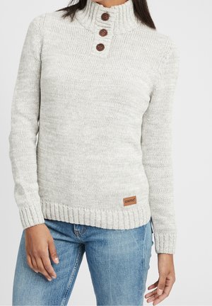 Femme portant un pull en tricot gris clair avec un col haut et trois boutons marron, associé à un jean bleu.