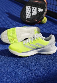 Bright yellow and white sports shoes on a blue court with a padel racket and ball near a net in the background.