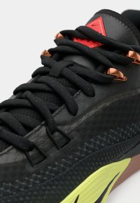 Κοντινός όψη μαύρου Sneakers με μαύρες κορδόνες, κόκκινο λογότυπο, χαλκούς συνδέσμους και φωτεινή κίτρινη λεπτομέρεια στη σόλα με σχέδιο μαύρου Swoosh.