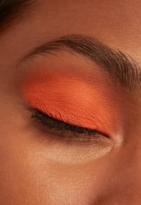 Gesloten oog met gladde bruine huid, gedurfde oranje oogschaduw, natuurlijke dikke wenkbrauw en gekrulde zwarte wimpers zichtbaar.