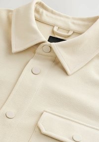 Beige košile na knoflíky s texturovanou látkou, s přeloženým límcem, třemi kulatými knoflíky a náprsní kapsou vepředu.